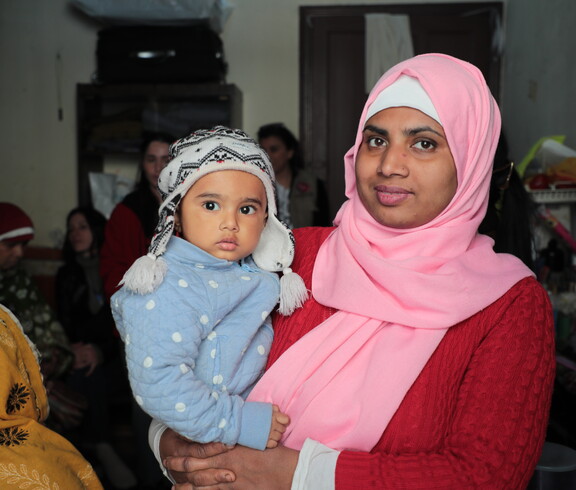Frau mit rosa Kopftuch hält ein kleines Kind im Arm und schaut in die Kamera.