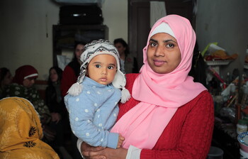Frau mit rosa Kopftuch hält ein kleines Kind im Arm und schaut in die Kamera.