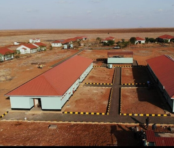 Luftaufnahme des neu errichteten Schulkomplexes der Tiigo School in Burgabo mit mehreren hellblauen Gebäuden unter roten Dächern, gepflasterten Wegen und schwarz-gelb markierten Gehsteigen inmitten der trockenen, roten Landschaft von Marsabit County, Kenia.