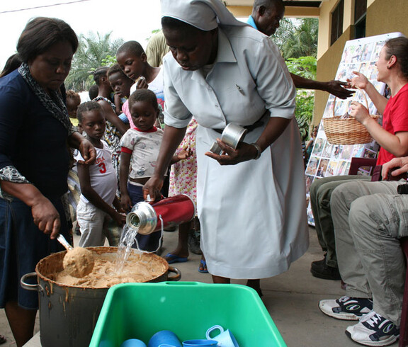 Krankenschwester gießt heißes Wasser in einen großen Topf mit Brei, umgeben von Kindern und einer weiteren Helferin in der Ernährungsstation in N’sele
