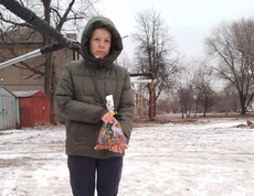 Eine Person in winterlicher Kleidung steht mit zwei Plastiktüten vor sich draußen. Die Landschaft ist schneebedeckt