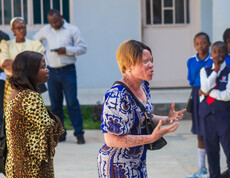 Eine Trainerin von FLEM spricht gestikulierend zu einer Gruppe; im Hintergrund stehen Erwachsene sowie Schülerinnen und Schüler in Schuluniform, die aufmerksam zuhören.