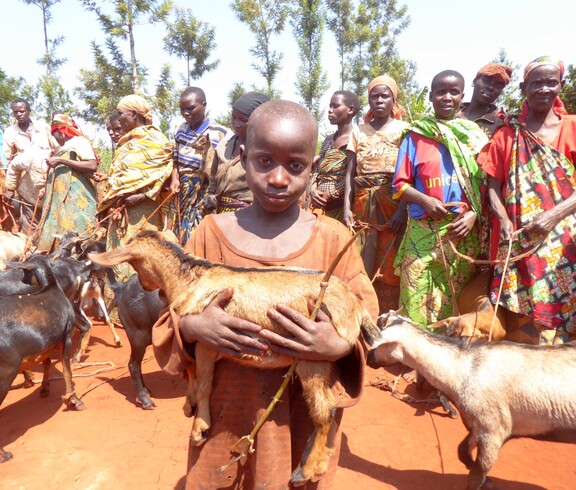Ein burundisches Kind steht vor einer Gruppe Frauen und trägt eine Ziege im Arm. 