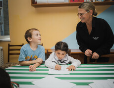 Zwei kleine Kinder grinsen ihre Betreuerin beim Hausaufgaben machen an.