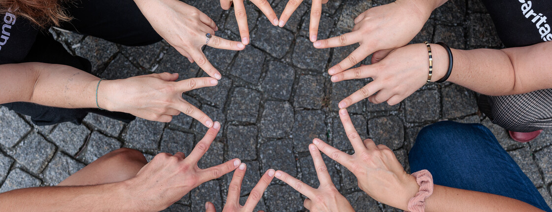 Fünf Personen sitzen im Kreis in der Hocke und formen mit den Zeige- und Mittelfingern einen Stern.