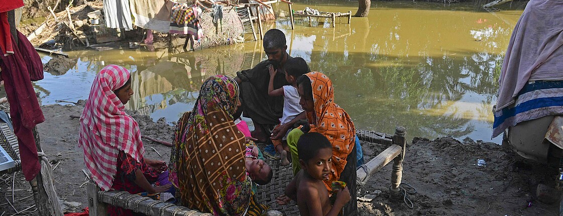 Eine Familie sitzt auf einem Bettgestell, im Hintergrund steht das Wasser von einer Überschwemmung, Reste eines zerstörten Hauses sind zu sehen.