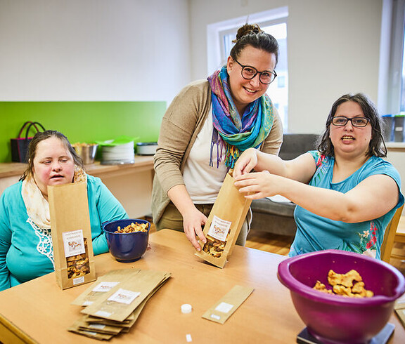Zwei Frauen mit Behinderung verpacken Lebensmittel an einem Tisch, begleitet von einer Betreuerin