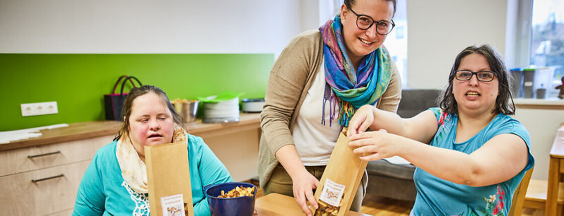 Zwei Frauen mit Behinderung verpacken Lebensmittel an einem Tisch, begleitet von einer Betreuerin