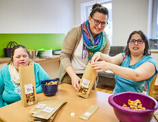 Zwei Frauen mit Behinderung verpacken Lebensmittel an einem Tisch, begleitet von einer Betreuerin