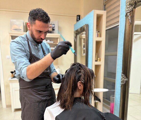 Eine Person führt eine Haarfärbung an einer sitzenden Person durch. Im Vordergrund sind eine Farbschale und Arbeitsutensilien zu sehen.