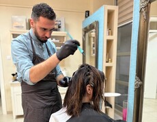 Eine Person führt eine Haarfärbung an einer sitzenden Person durch. Im Vordergrund sind eine Farbschale und Arbeitsutensilien zu sehen.