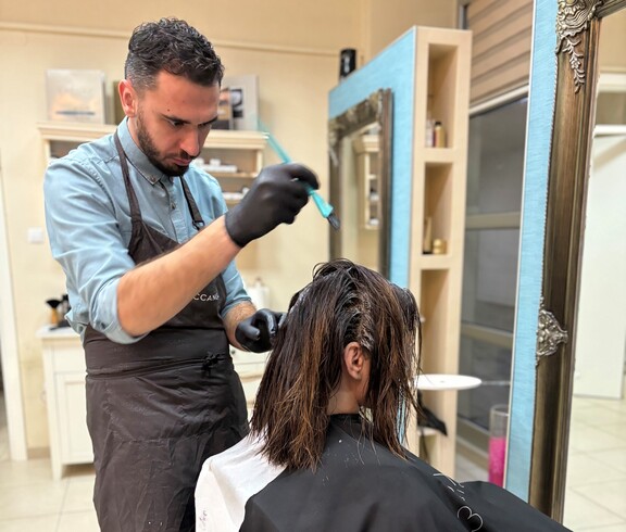 Eine Person führt eine Haarfärbung an einer sitzenden Person durch. Im Vordergrund sind eine Farbschale und Arbeitsutensilien zu sehen.