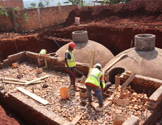 Zwei Bauarbeiter in gelben Warnwesten und Schutzhelmen arbeiten auf einer Baustelle mit gewölbten Betonkuppeln und Ziegelmauerwerk – Errichtung einer Biogasanlage im Rahmen des Caritas-Klimaprojekts in Burundi.