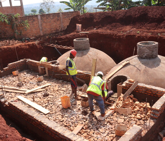 Zwei Bauarbeiter in gelben Warnwesten und Schutzhelmen arbeiten auf einer Baustelle mit gewölbten Betonkuppeln und Ziegelmauerwerk – Errichtung einer Biogasanlage im Rahmen des Caritas-Klimaprojekts in Burundi.