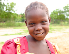Nahaufnahme eines lächelnden Mädchens in rotem Kleid auf dem Schulgelände in Gumbo, Südsudan.