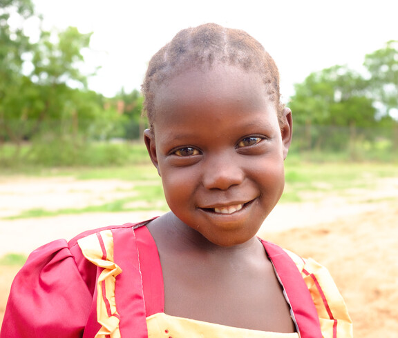 Nahaufnahme eines lächelnden Mädchens in rotem Kleid auf dem Schulgelände in Gumbo, Südsudan.