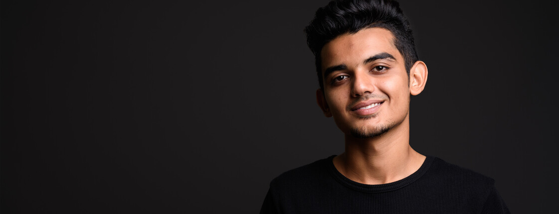 Ein junger Mann lächelt leicht in die Kamera, er trägt ein schwarzes Shirt. Der Hintergrund ist dunkel.