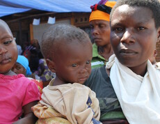 Eine Mutter hält ihr kleines Kind im Arm und blickt ernst in die Kamera; das Kind wirkt erschöpft und untergewichtig – Szene vor dem Ernährungszentrum in Gitega, Burundi, im Hintergrund weitere wartende Mütter.