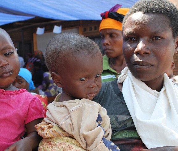 Eine Mutter hält ihr kleines Kind im Arm und blickt ernst in die Kamera; das Kind wirkt erschöpft und untergewichtig – Szene vor dem Ernährungszentrum in Gitega, Burundi, im Hintergrund weitere wartende Mütter.