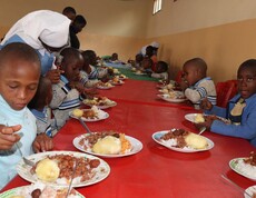Burundische Kinder sitzen an einem langen Tisch mit rotem Tischtuch und haben einen Teller Essen vor sich. Es gibt Maisbrei und Bohnen. 