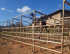 In Lubumbashi entsteht ein Solartrockner für die umweltfreundliche Trocknung von Ernten  Zwei Personen bauen eine große Holzkonstruktion auf einem trockenen Feld auf.