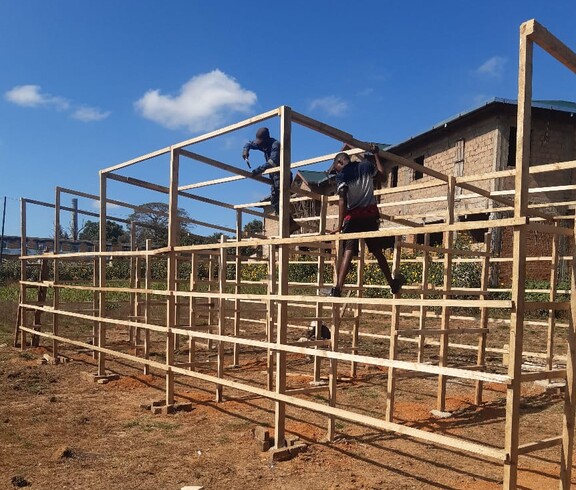 In Lubumbashi entsteht ein Solartrockner für die umweltfreundliche Trocknung von Ernten  Zwei Personen bauen eine große Holzkonstruktion auf einem trockenen Feld auf.