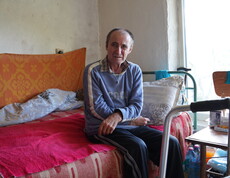 Ein älterer Mann sitzt auf einem Sofabett und blickt in die Kamera.