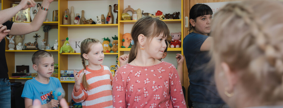 Kinder stehen in einem Raum und tanzen. Im Hintergrund sind Regale mit Spielsachen und Deko-Gegenständen zu sehen.