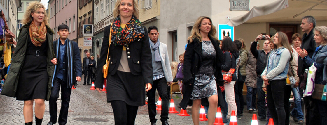 Eine Modenschau, mitten in einer Stadt. Frauen und Männer präsentieren ihre Kleidung.