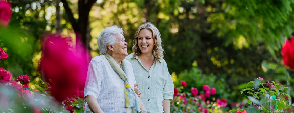Eine junge Frau begleitet eine Seniorin mit Rollator bei einem Spaziergang durch einen Rosengarten.