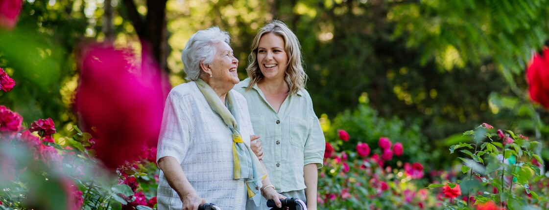 Eine junge Frau begleitet eine Seniorin mit Rollator bei einem Spaziergang durch einen Rosengarten.