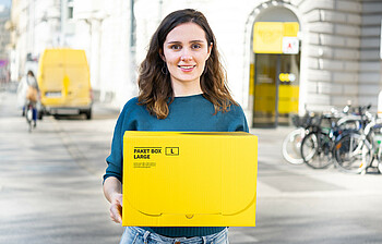 Junge Frau hält ein gelbes Postpaket, sie lächelt und schaut in die Kamera. Im Hintergrund ist eine Postfiliale.