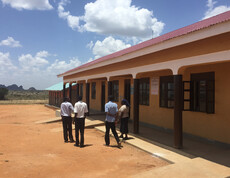Ein langes, gelbes Schulgebäude mit geöffneten Fenstern, davor vier Personen, die über den Schulhof gehen.