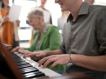 Eine junger Mann und eine Seniorin spielen gemeinsam Klavier.
