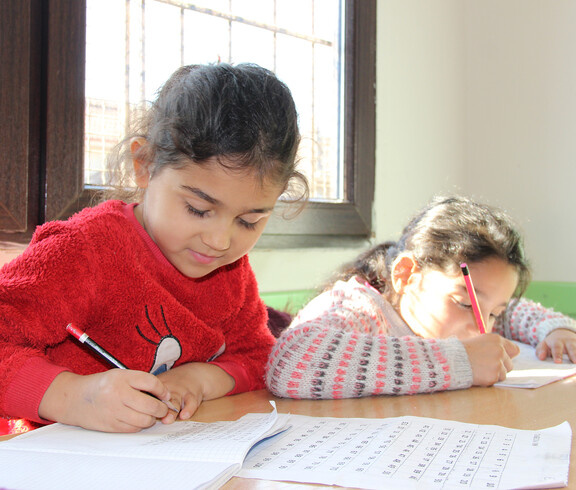 Zwei Mädchen sitzen an einem Tisch und schreiben konzentriert in ihre Hefte. Sonnenlicht fällt durch ein Fenster auf die Tischfläche, beide wirken ruhig und fokussiert.
