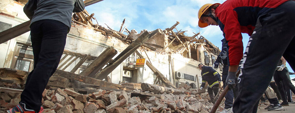 Männer stehen vor eingestürzten Gebäuden und schaufeln Trümmer weg