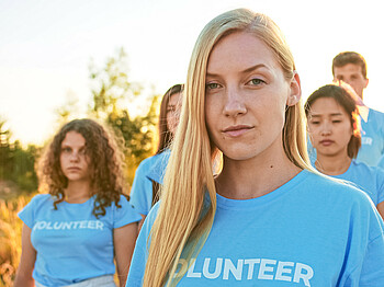 Portrait einer jungen Frau in blauem T-Shirt mit mehreren Personen im Hintergrund.