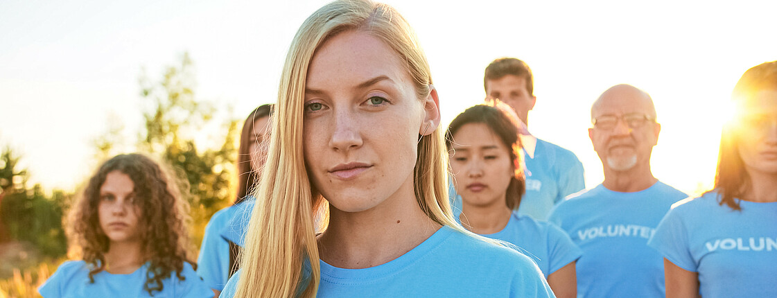 Portrait einer jungen Frau in blauem T-Shirt mit mehreren Personen im Hintergrund.