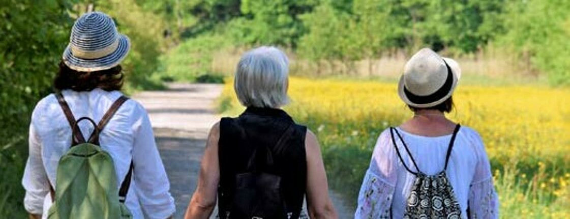 Drei Frauen mit Rucksäcken gehen Hand in Hand einen Feldweg.