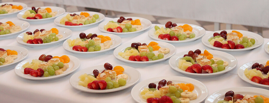 Ein Tisch mit weißer Tischdecke, auf dem viele Teller mit Früchten aufgedeckt sind.