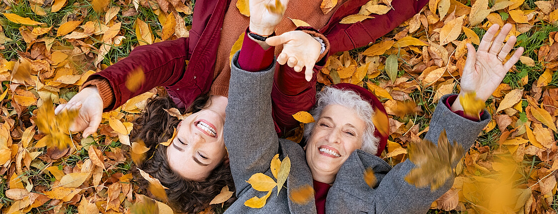 Eine ältere und eine jüngere Frau liegen auf dem Rücken am Boden und werfen buntes Herbstlaub in die Luft.