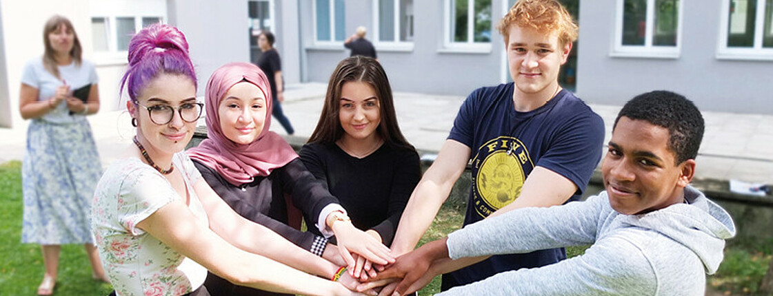 3 Schülerinnen und 2 Schüler stehen im Halbkreis beieinander im Grünen im Hof der Schule und Halten die Hände übereinander. Im Hintergrund steht verschwommen eine Schülerin mit Buch in der Hand. Copyright Caritas Schule Wiener Neustadt