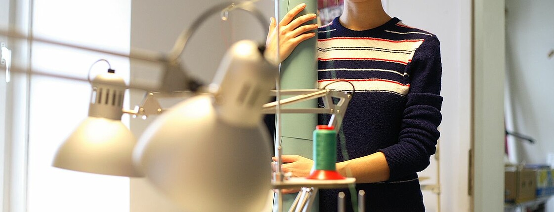 Eine junge Frau steht in einer Stoff-Werkstatt und hält sich an einer Papierrolle fest.