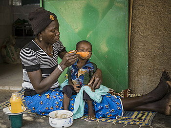Eine Mutter sitzt auf dem Boden und füttert ihr Kind, welches auf ihrem Schoss sitzt.