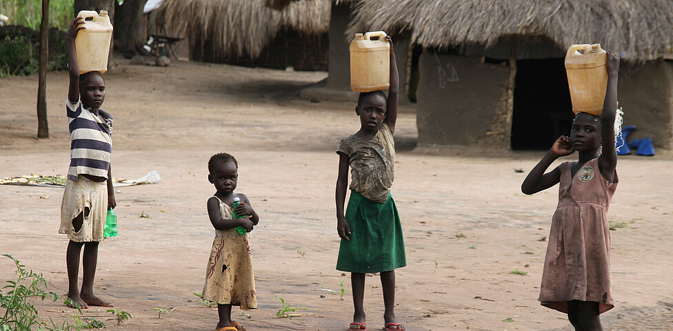 Drei Kinder tragen einen gelben Wasserkanister am Kopf und sehen in die Kamera. Ein weiteres Kind hat eine Wasserflasche in der Hand.
