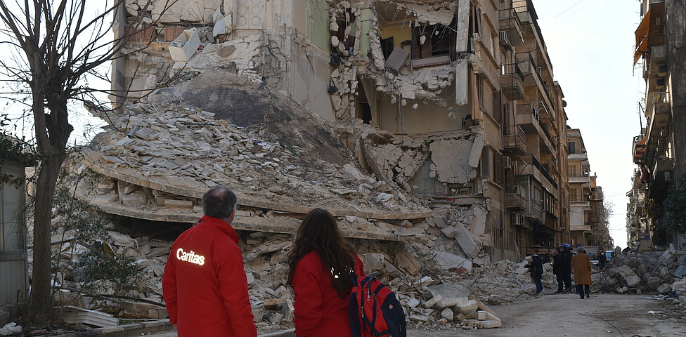 zwei Mitarbeiter*innen der Caritas Österreich stehen vor eingestürzten Gebäuden in Aleppo