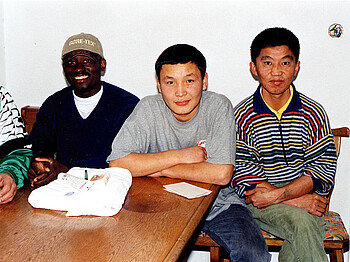 Drei Männer (ein Afrikaner, ein Asiate und ein Süd-Amerikaner) sitzen an einem Tisch nebeneinander uns schauen in die Kamera.