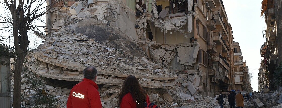 Es sind zwei Personen mit Caritas-Jacken von hinten zu sehen, sie stehen vor Trümmern eines Hauses.