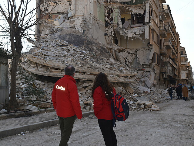 Es sind zwei Personen mit Caritas-Jacken von hinten zu sehen, sie stehen vor Trümmern eines Hauses.