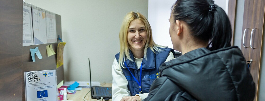 Two women talking to eachother in an office booth, one wearing Caritas staff clothing. She is smiling and holding the hands of the other woman. 